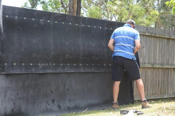 A man in a blue striped shirt installing an acoustic fence wrap on his backyard fence